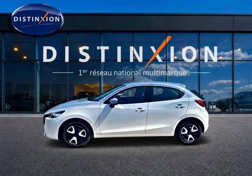 Vue de profil côté gauche d'une Mazda2 blanche 2024 garée devant une vitrine avec logo Distinxion.