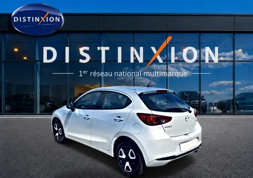 Vue 3/4 arrière droite d'une Mazda2 blanche 2024 avec jantes distinctives et vitres teintées, devant une vitrine Distinxion.