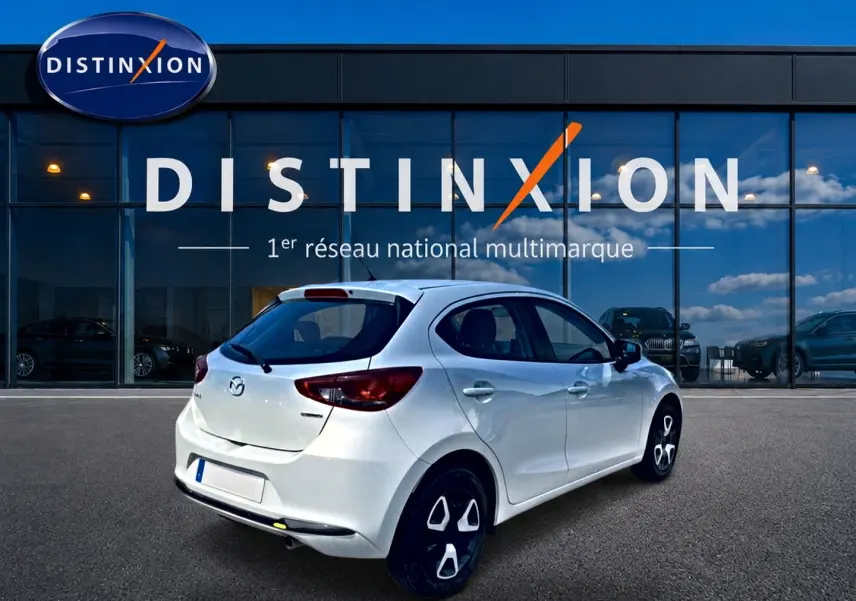 Vue 3/4 arrière droite d'une Mazda2 blanche 2024 stationnée devant une vitrine Distinxion.