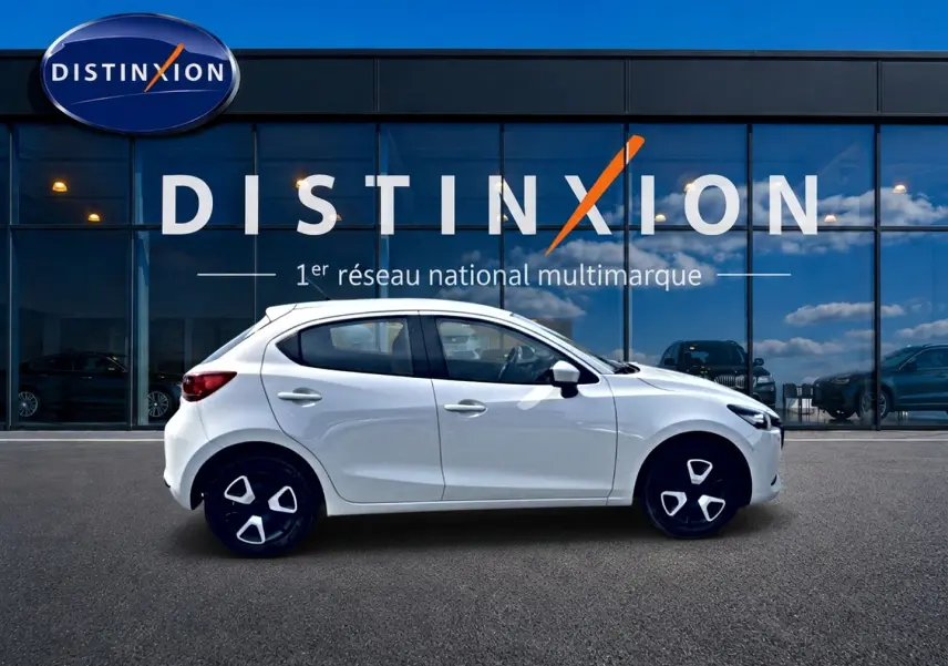 Vue latérale droite d'une Mazda2 blanche 2024 avec jantes noires et détails modernes, stationnée devant une vitrine Distinxion.