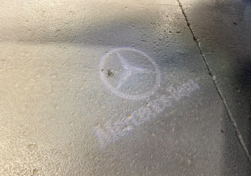 Projection lumineuse au sol du logo Mercedes-Benz en blanc sur un sol gris texturé, sans véhicule visible.