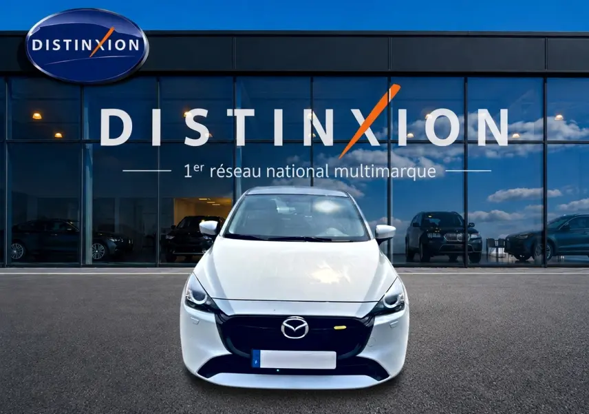 Vue avant d'une Mazda2 blanche 2024 avec calandre noire, stationnée devant une vitrine Distinxion.