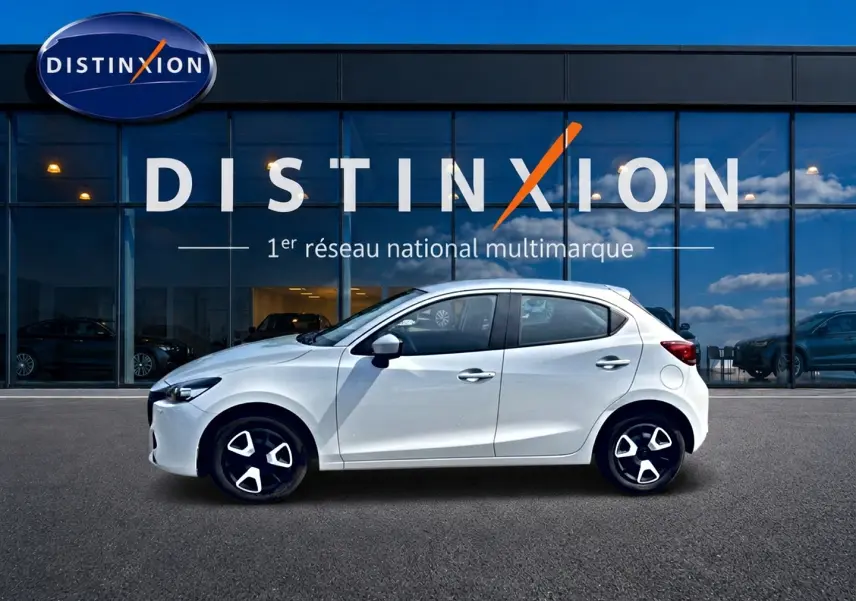 Vue latérale droite d'une Mazda2 blanche 2024 garée devant une vitrine avec logo Distinxion.