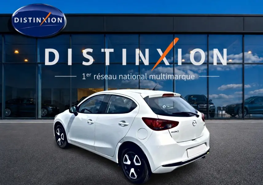 Vue 3/4 arrière droite d'une Mazda2 blanche 2024 avec jantes distinctives, garée devant une vitrine Distinxion.
