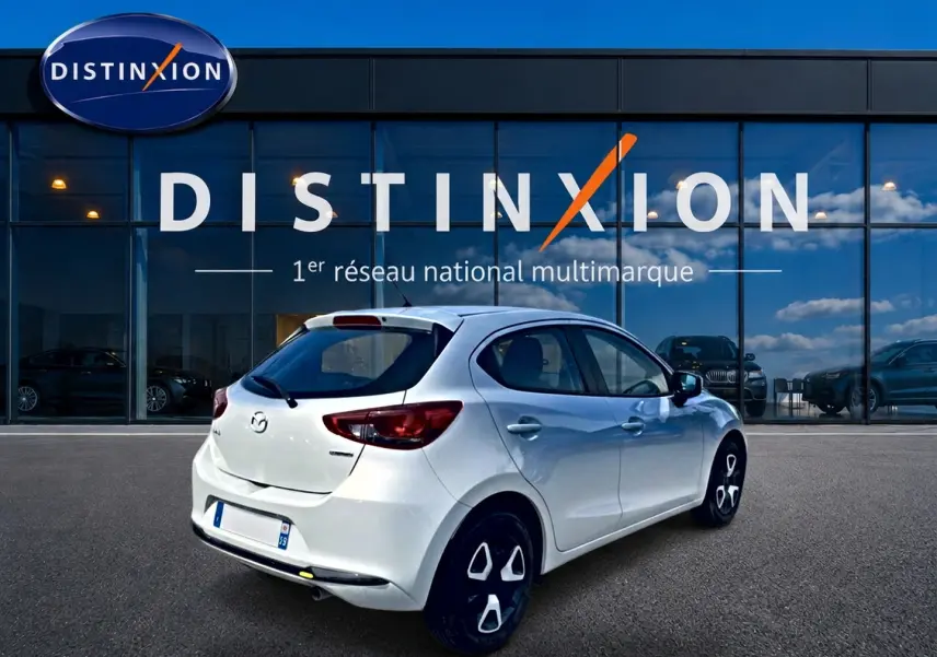 Vue 3/4 arrière droite d'une Mazda2 blanche 2024 garée devant une vitrine avec logo Distinxion.