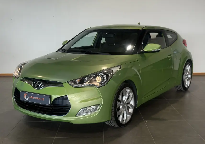 Hyundai Veloster vert 2025 en 3/4 avant droit, avec phares allumés et jantes alliage visibles.