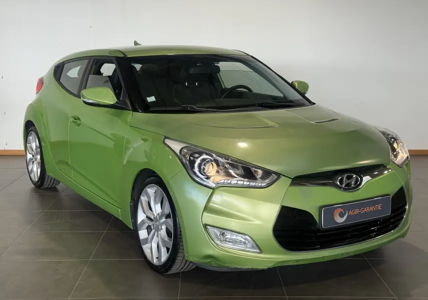 Vue 3/4 avant d'une Hyundai Veloster 2025 vert métallisé avec phares allumés et jantes argentées.