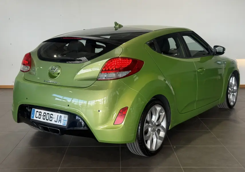 Vue 3/4 arrière droite d'une Hyundai Veloster 2025 vert métallisé avec toit noir et feux arrière allumés.