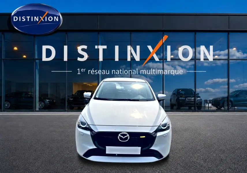 Vue avant d'une Mazda2 blanche 2024 stationnée devant une vitrine avec logo Distinxion en arrière-plan.