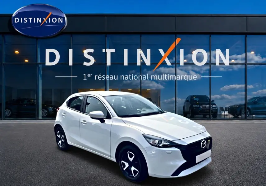 Vue 3/4 avant droite d'une Mazda2 blanche 2024 avec jantes noires et calandre noire devant un showroom Distinxion.