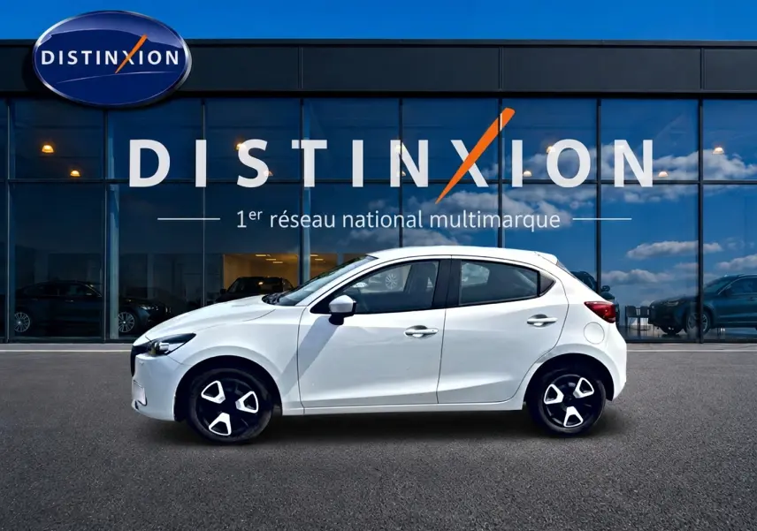 Vue de profil côté gauche d'une Mazda2 blanche 2024 garée devant une vitrine avec logo Distinxion.