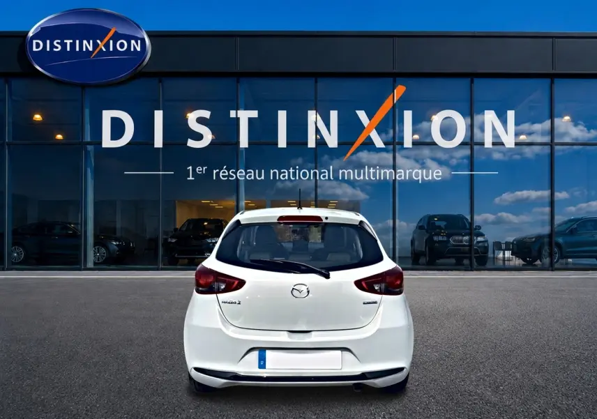 Vue arrière d'une Mazda2 blanche 2024 garée devant une vitrine avec logo Distinxion en arrière-plan.