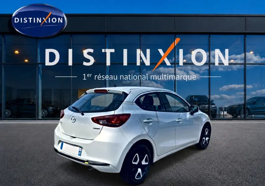 Vue 3/4 arrière droite d'une Mazda2 blanche 2024 stationnée devant une vitrine Distinxion.
