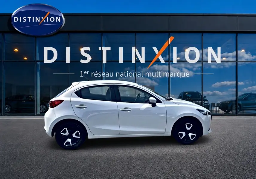 Vue latérale droite d'une Mazda2 blanche 2024 garée devant une vitrine avec logo Distinxion.