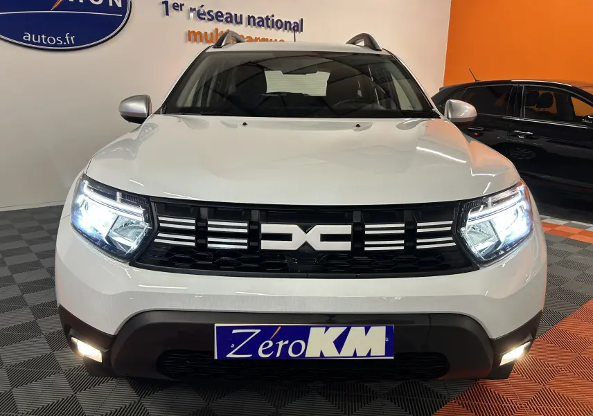Vue avant d'un Dacia Duster blanc 2025 avec phares allumés et plaque Zéro KM en intérieur showroom.