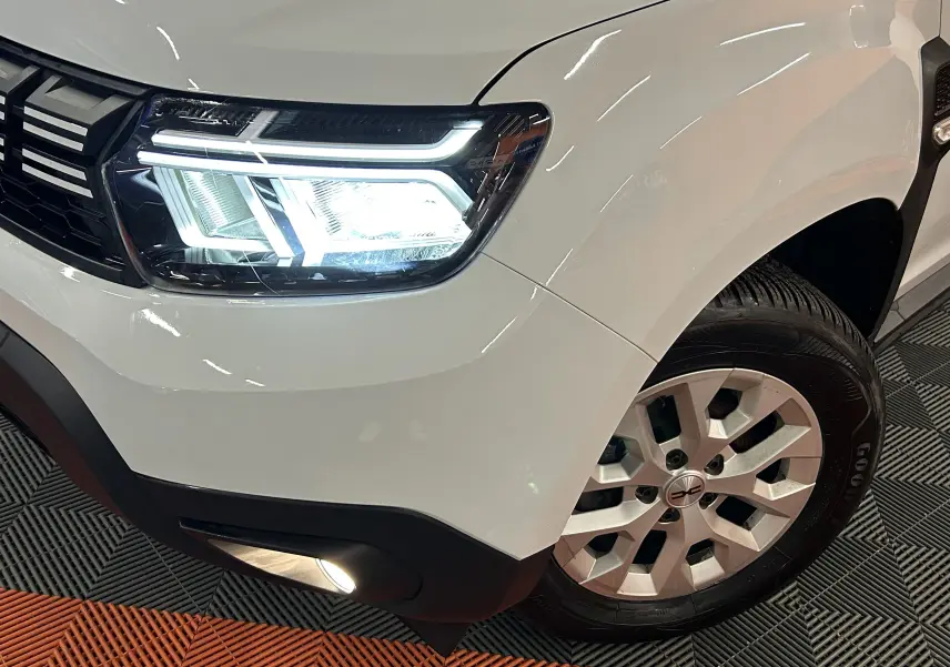 Gros plan sur l'avant droit blanc du Dacia Duster 2025 avec phare LED allumé et jante alu distinctive.
