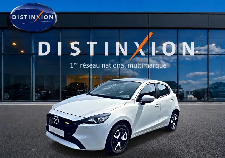 Vue 3/4 avant droite d'une Mazda2 blanche 2024 avec calandre noire et jantes distinctives devant un showroom Distinxion.