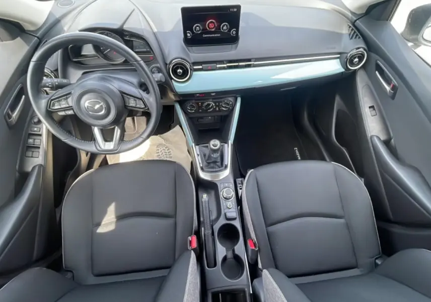 Vue intérieure de la Mazda2 2024 montrant le tableau de bord, volant cuir multifonction et console centrale avec levier manuel.