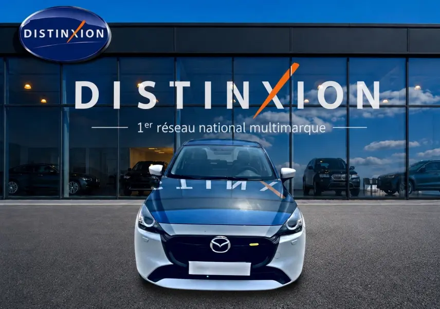 Vue avant d'une Mazda2 blanche 2024 avec calandre noire, stationnée devant une vitrine de concession multimarque.