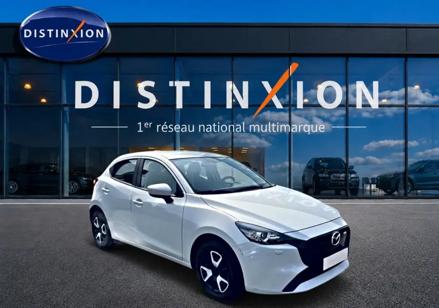 Vue 3/4 avant gauche d'une Mazda2 blanche 2024 avec calandre noire et jantes distinctives devant un showroom Distinxion.