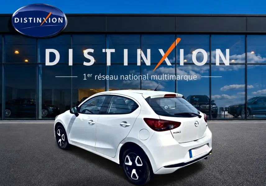 Vue 3/4 arrière droite d'une Mazda2 blanche 2024 avec jantes noires et détails modernes, devant une vitrine Distinxion.