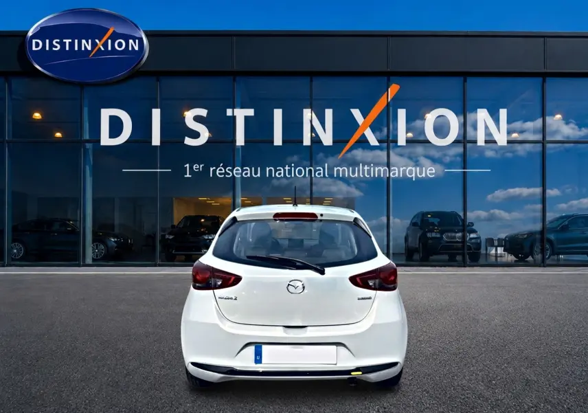 Vue arrière d'une Mazda2 blanche 2024 garée devant une vitrine avec logo Distinxion en arrière-plan.