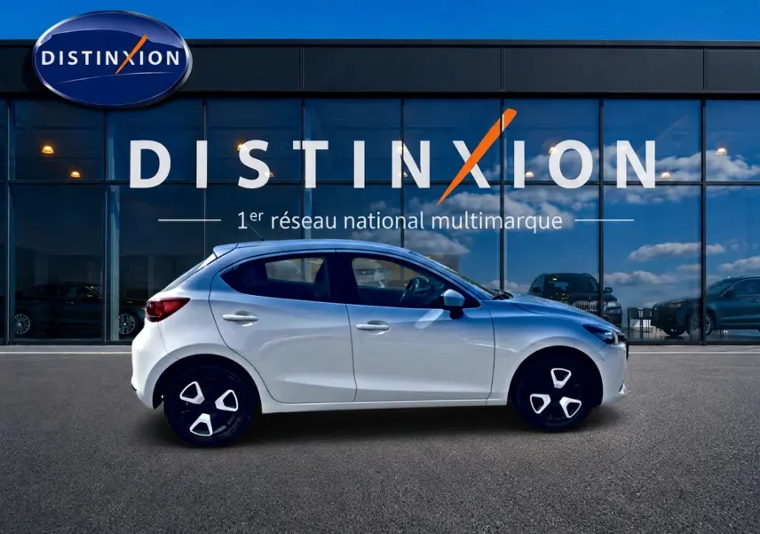 Vue de profil côté gauche d'une Mazda2 blanche 2024 garée devant une vitrine avec logo Distinxion.