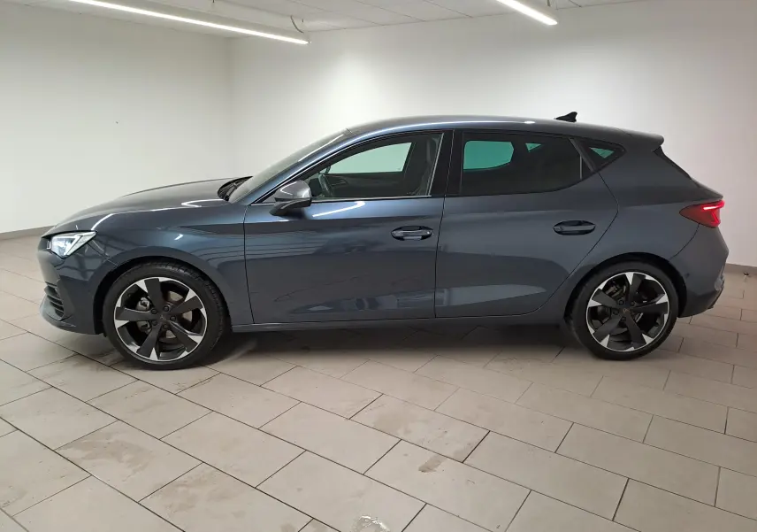 Vue latérale droite d'une CUPRA Leon 2023 gris magnétique avec jantes noires et feux arrière allumés dans un intérieur neutre.