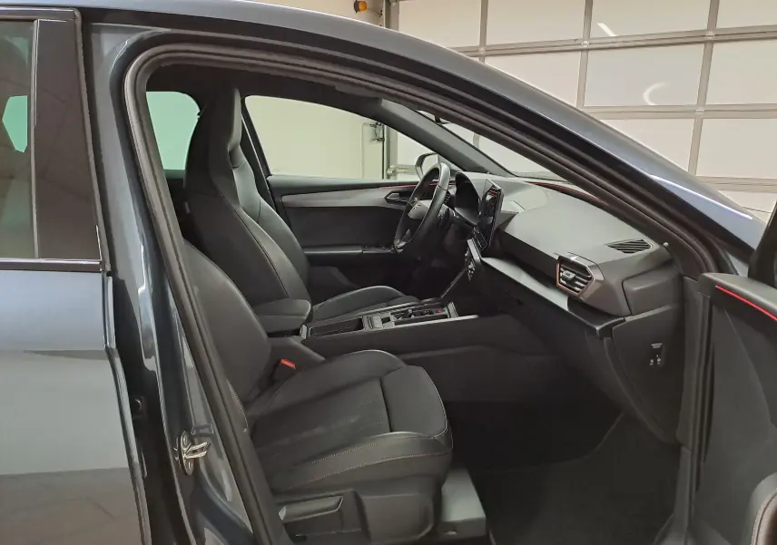 Vue intérieure côté gauche de la CUPRA Leon 1.5 TSI 150 DS7 V 2023 en gris magnétique, sièges noirs avec surpiqûres orange.