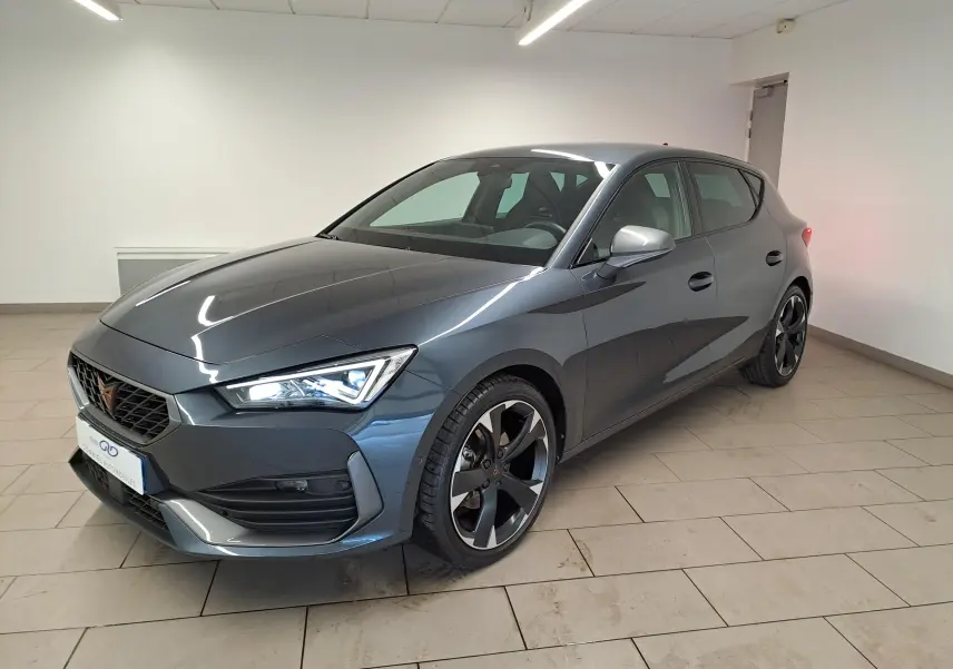 Vue 3/4 avant d'une CUPRA Leon 1.5 TSI 150 DS7 V gris magnétique avec jantes noires et phares allumés en intérieur.