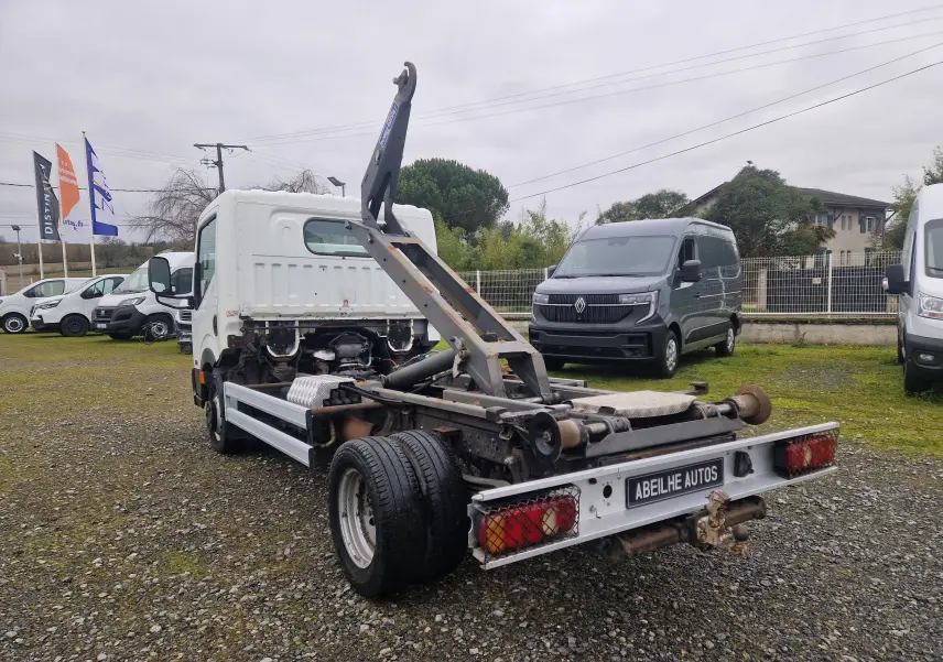 Vue 3/4 arrière droite d'un Nissan Cabstar blanc 2010 avec châssis cabine et bras polybenne visible sur terrain gravillonné.