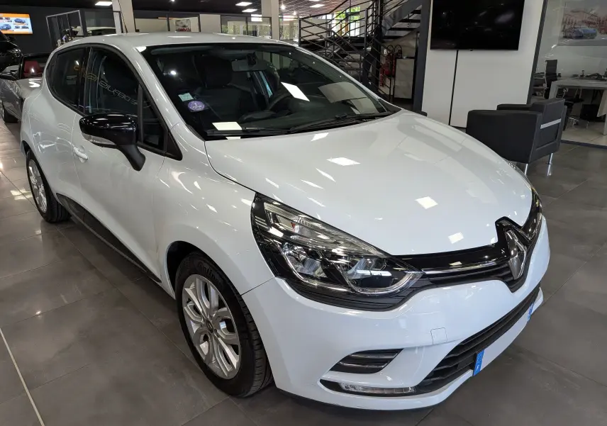 Vue 3/4 avant d'une Renault Clio IV TCe 75 blanc glacier, avec rétroviseurs noirs et jantes alliage 16 pouces en intérieur showroom.