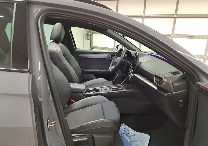 Vue intérieure côté conducteur de la CUPRA LEON ST gris Graphène, avec sièges cuir noirs et éclairage d’ambiance bleu.