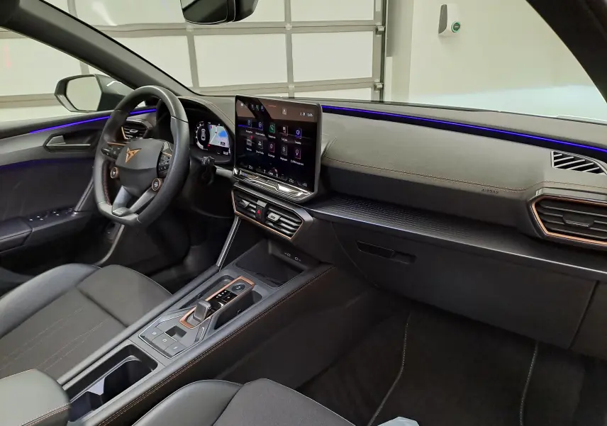 Vue intérieure côté conducteur de la CUPRA Leon ST 2025, tableau de bord moderne avec écran tactile et éclairage d’ambiance violet.