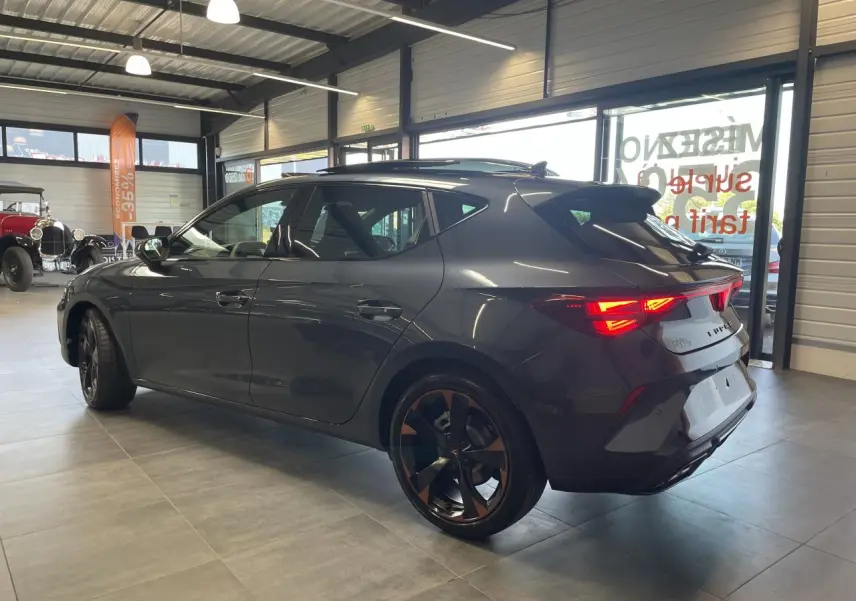 Vue 3/4 arrière droite d'une CUPRA Leon gris magnétique avec jantes cuivrées et feux arrière allumés en intérieur showroom.