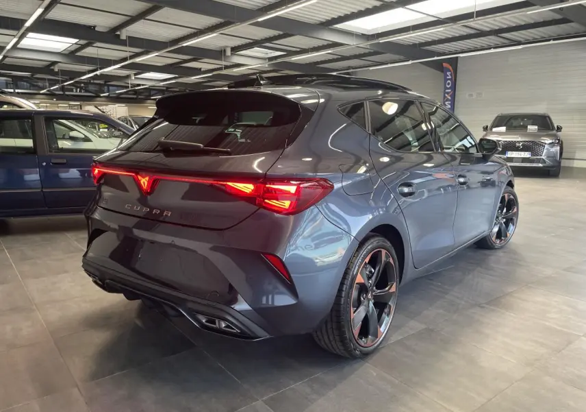 Vue 3/4 arrière droite d'une CUPRA Leon gris magnétique avec jantes cuivrées et feux arrière allumés en showroom.