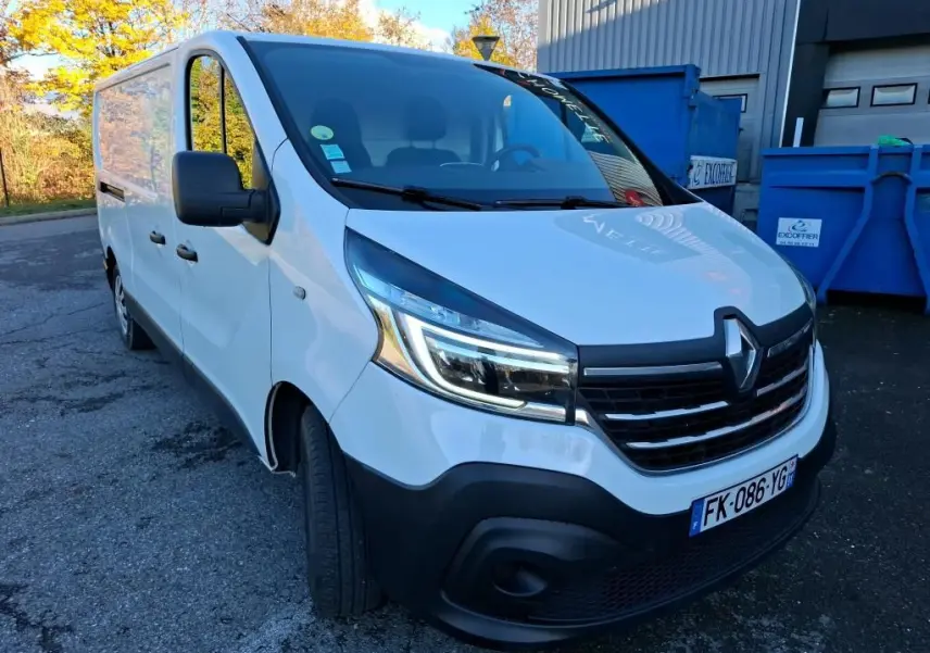 Vue 3/4 avant gauche d'un Renault Trafic Fourgon blanc de 2019 avec calandre noire et phares LED allumés.