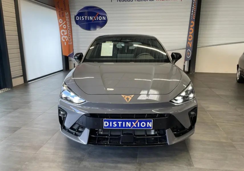 Vue avant d'une CUPRA Leon gris magnétique avec logo cuivré, phares allumés et calandre noire en showroom.