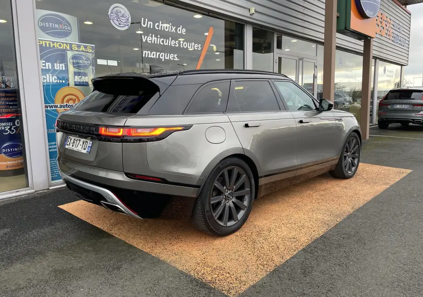 Vue 3/4 arrière droite d'un Range Rover Velar D300 gris Silicon Silver avec toit noir et jantes foncées, stationné devant un showroom.
