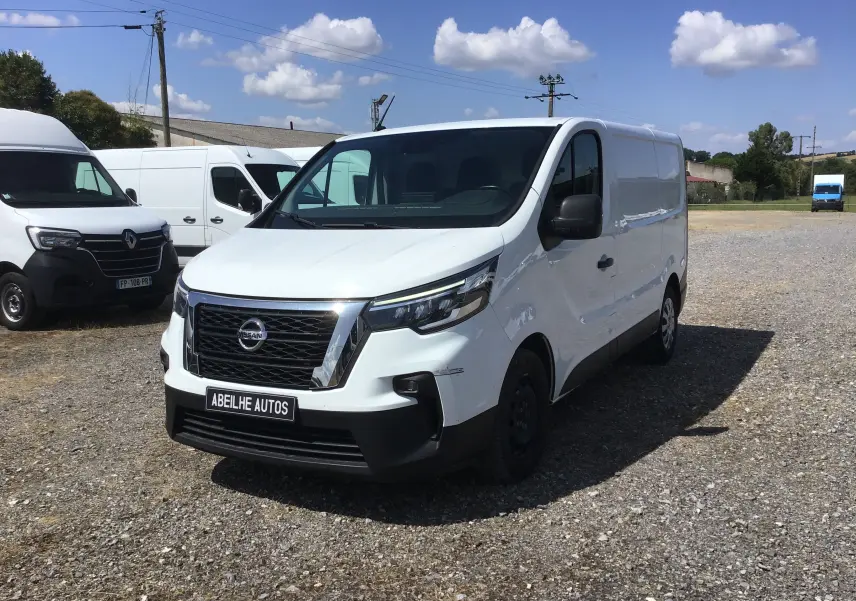Fourgon blanc Nissan Primastar L1H1 vu en 3/4 avant droit sur un terrain extérieur sous ciel bleu.