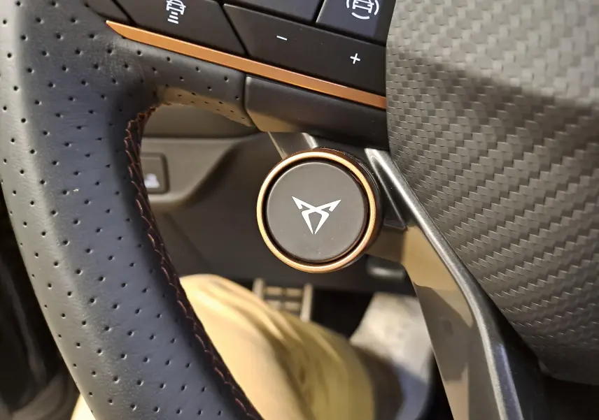 Gros plan sur le bouton rotatif du volant perforé avec logo CUPRA, entouré de finitions cuivre et carbone.