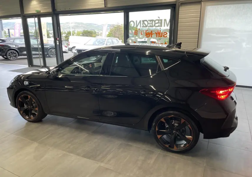 Vue latérale droite d'une CUPRA Leon noire avec jantes alliage 18'' cuivrées et feux arrière allumés en intérieur showroom.