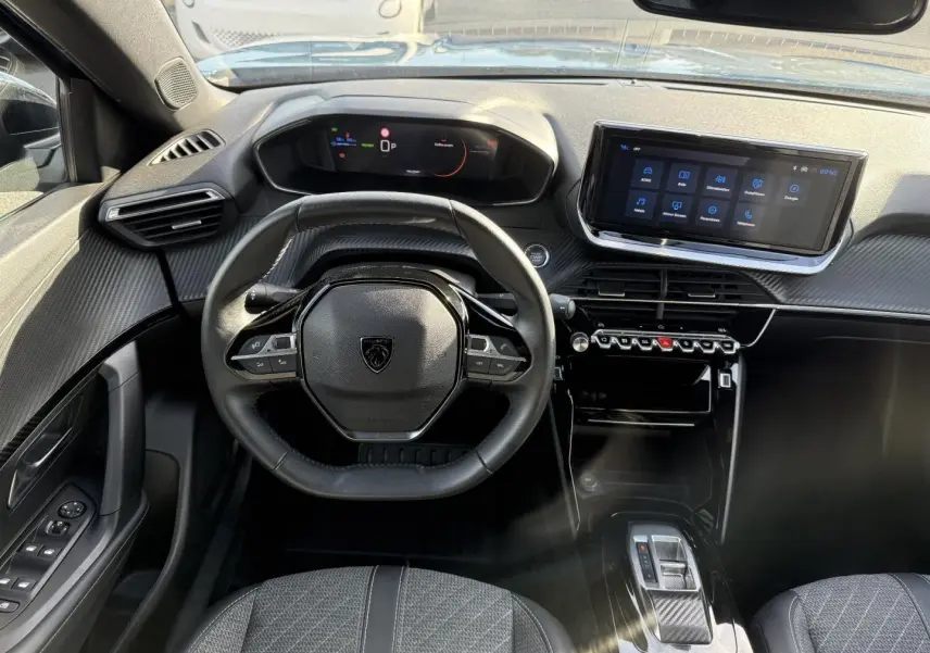 Vue intérieure avant du poste de conduite du Peugeot e-2008 2025 avec volant compact, écran tactile et finition carbone noire.