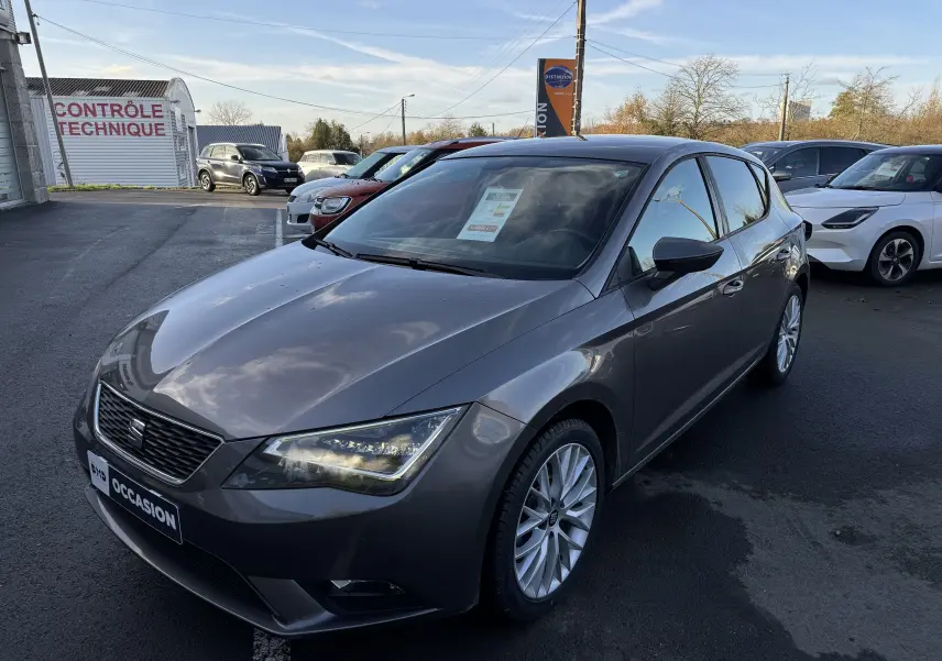 Vue 3/4 avant d'une SEAT Leon gris métallisé 2016 garée sur un parking avec phares allumés.