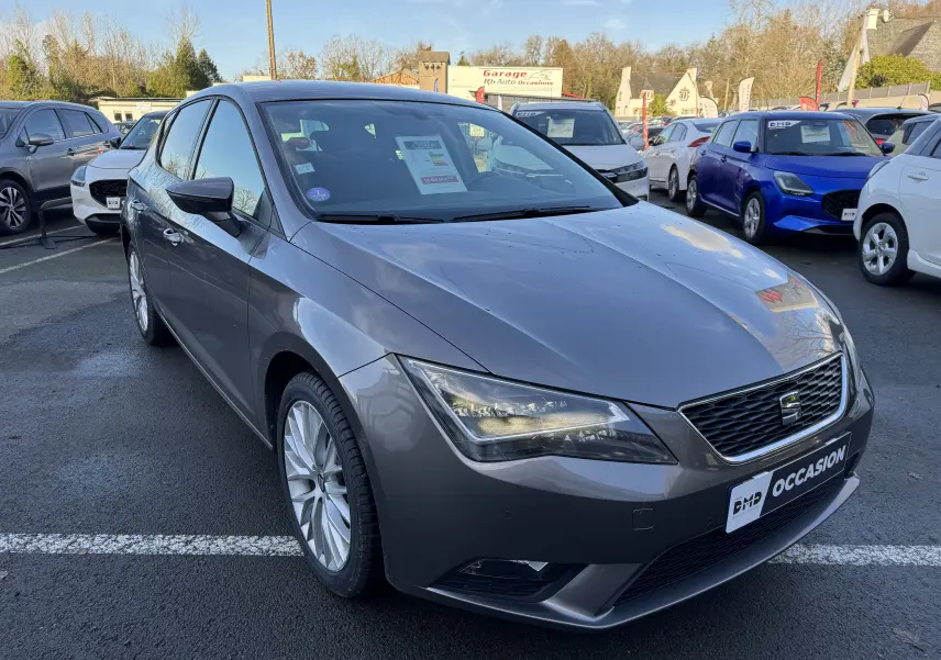 SEAT Leon gris vue 3/4 avant droit sur parking, avec phares allumés et jantes alliage visibles.