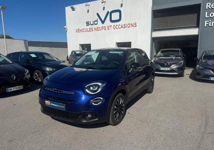Fiat 500X bleu vue 3/4 avant droit, avec phares ronds et jantes classiques 16 pouces, stationnée devant un garage.