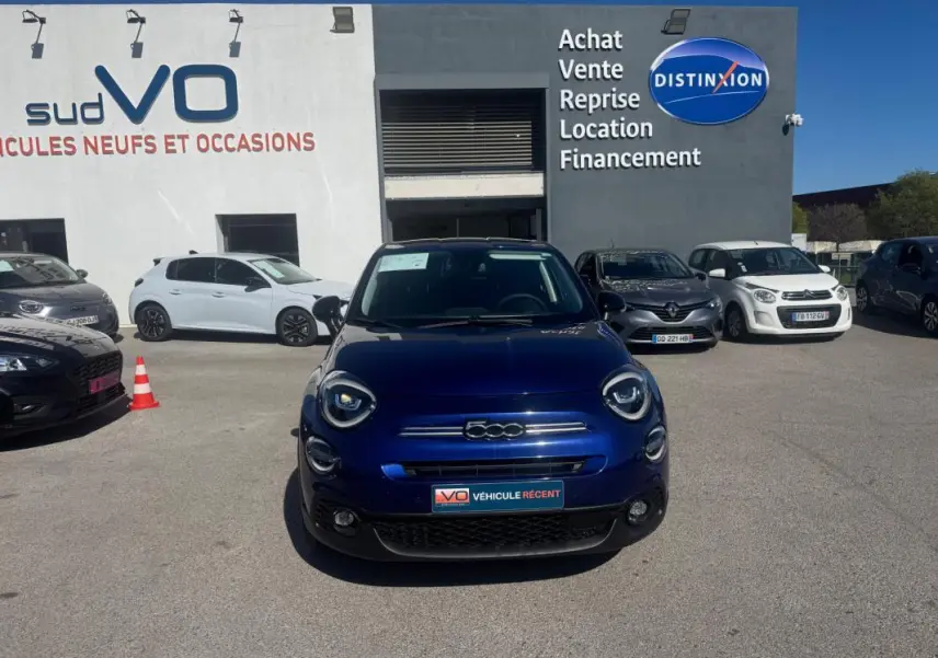 Vue de face d'une FIAT 500X bleu foncé stationnée devant un garage avec panneaux publicitaires visibles.