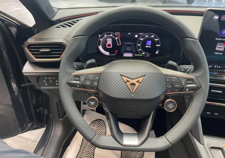 Vue intérieure centrée sur le volant perforé noir avec logo Cupra cuivré et tableau de bord digital couleur.