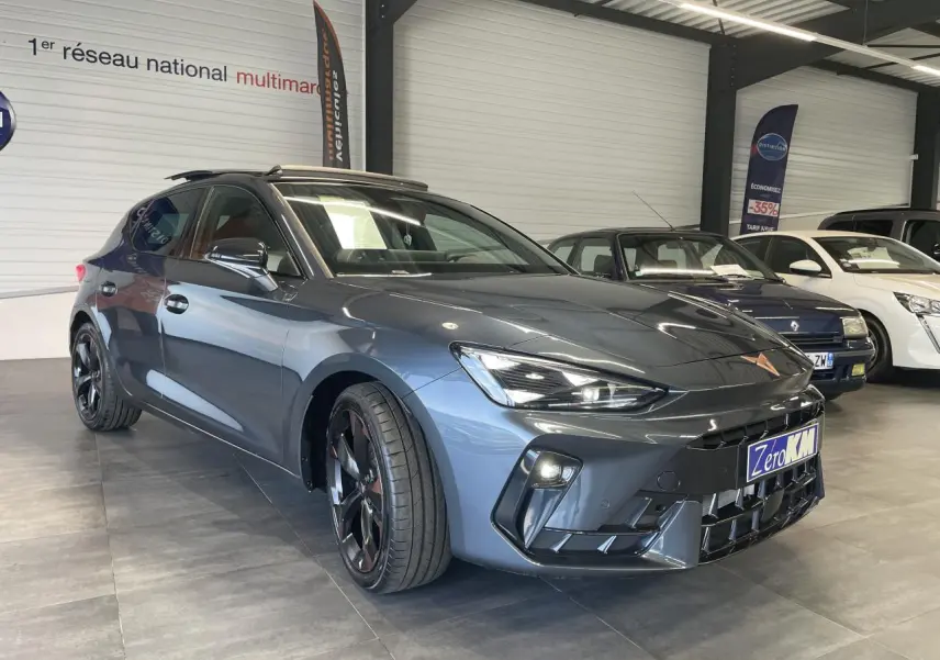 Vue 3/4 avant d'une CUPRA Leon gris magnétique avec jantes cuivrées et signature lumineuse Tri-LED en showroom.