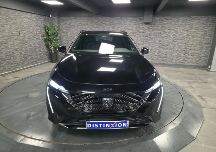 Vue avant d'une Peugeot 308 SW GT noire 2024 avec calandre noire brillante et feux LED allumés dans un showroom.